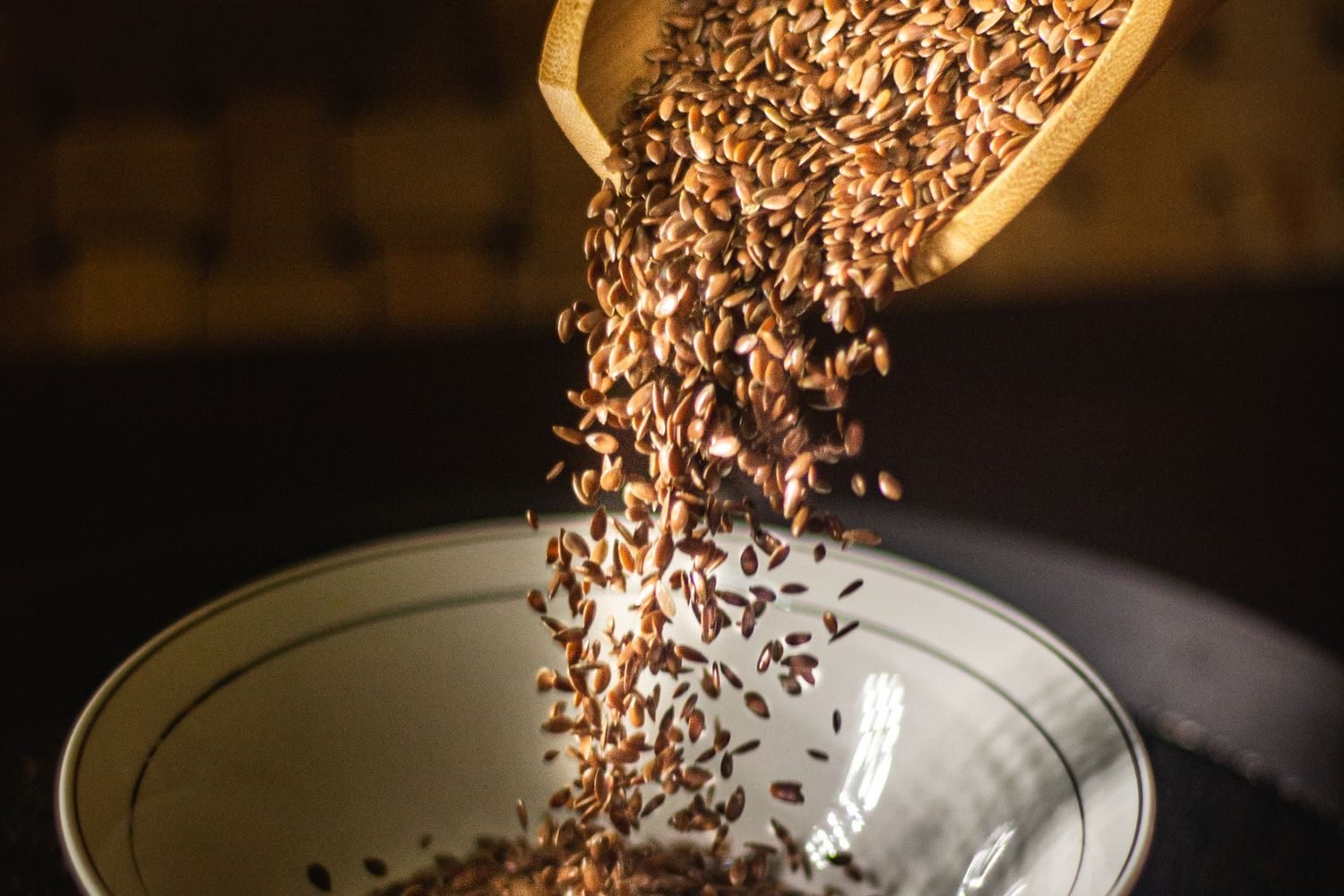 Ground flaxseed in a small bowl next to a spoon and flax seeds scattered on a kitchen counter